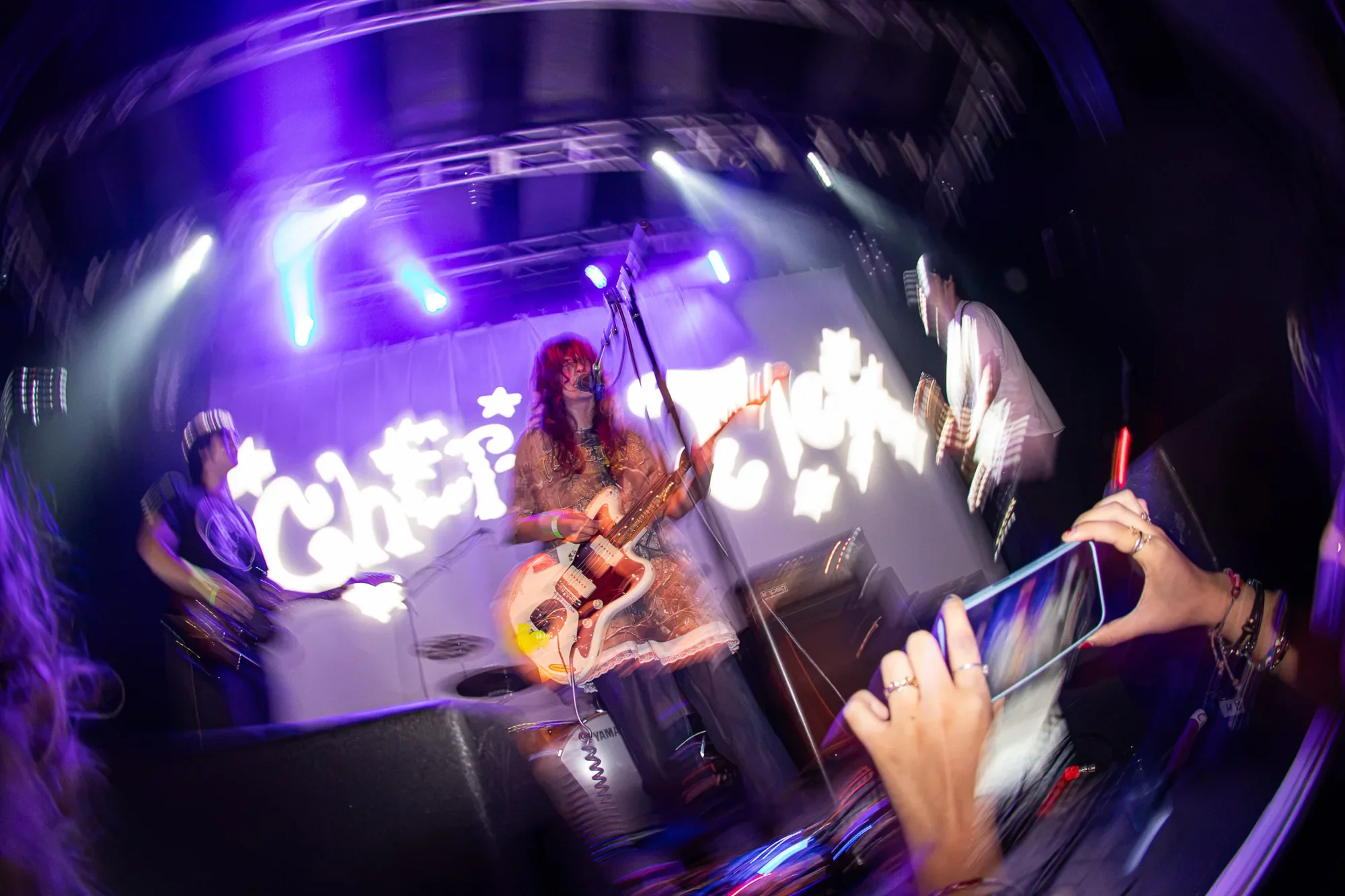 An image of Cherry Pick. Highlighting their frontman with her bright red hair lit up by the stage lights. It's a long exposure wide angle shot with the sides of the image blurred in a swirl.