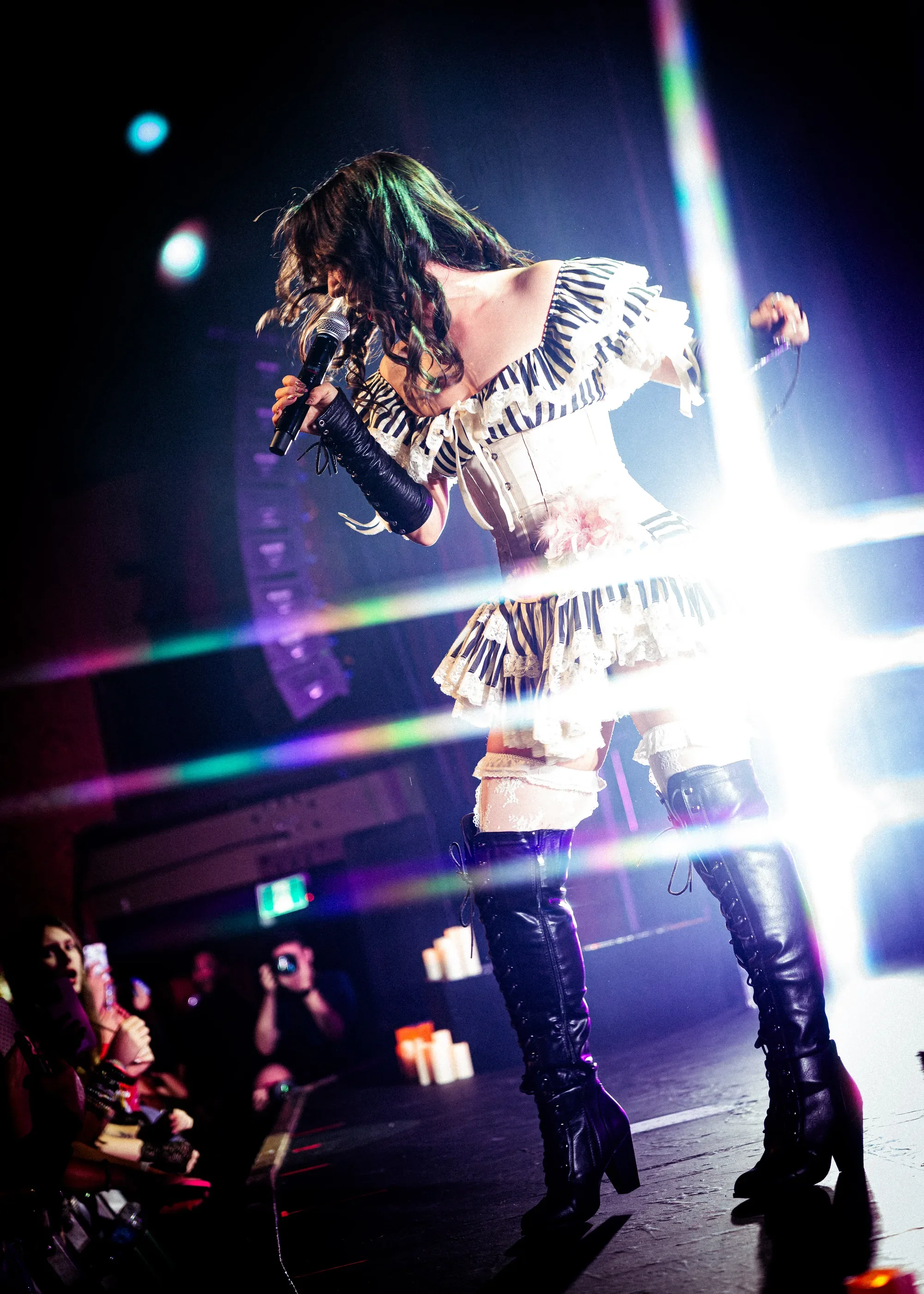 Emei performs at the edge of the stage in a striped, ruffled outfit and black thigh-high boots, with bright backlighting behind her.