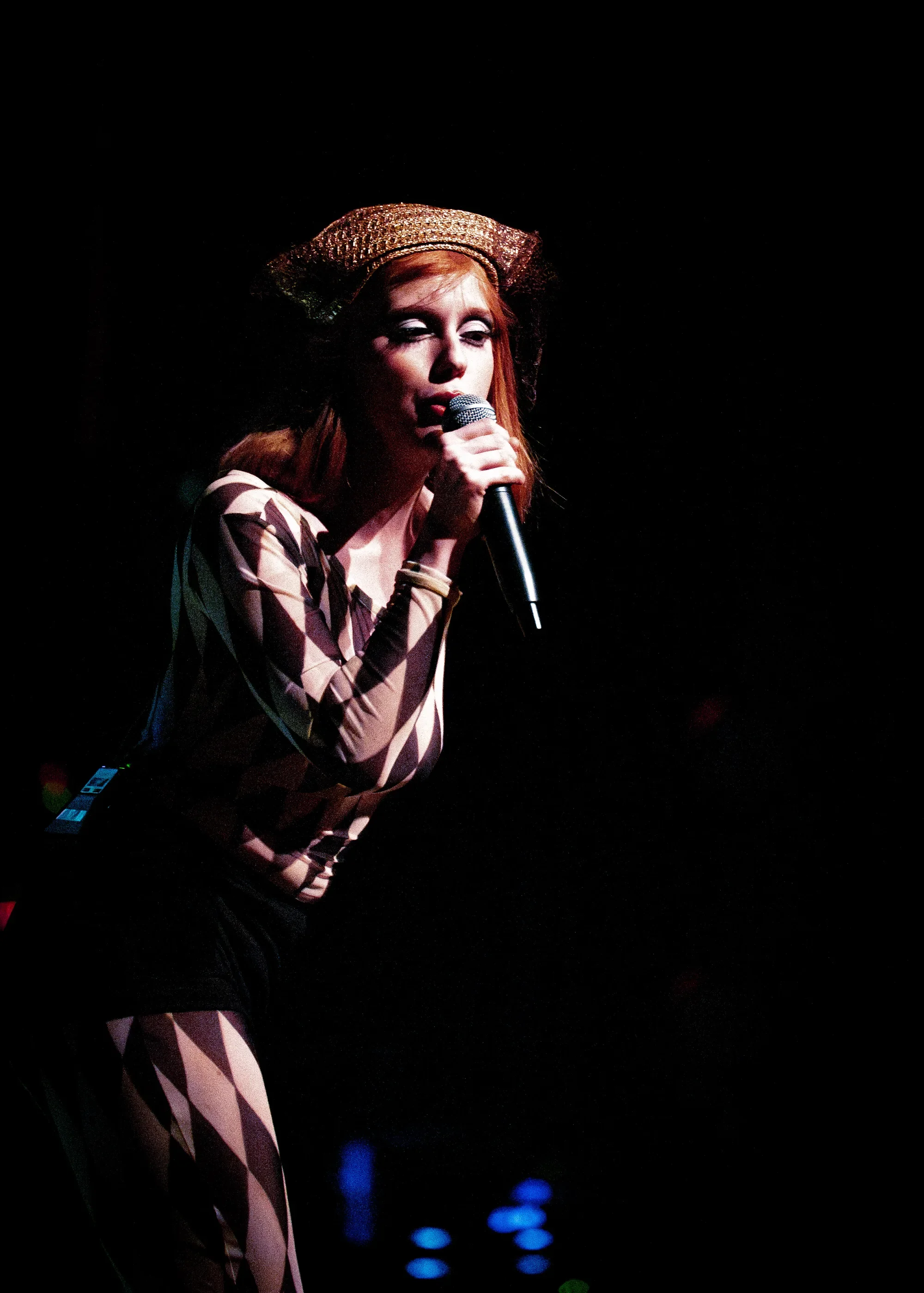Madelline leans forward to sing into a handheld microphone, wearing a diamond-patterned outfit and a gold-toned hat.