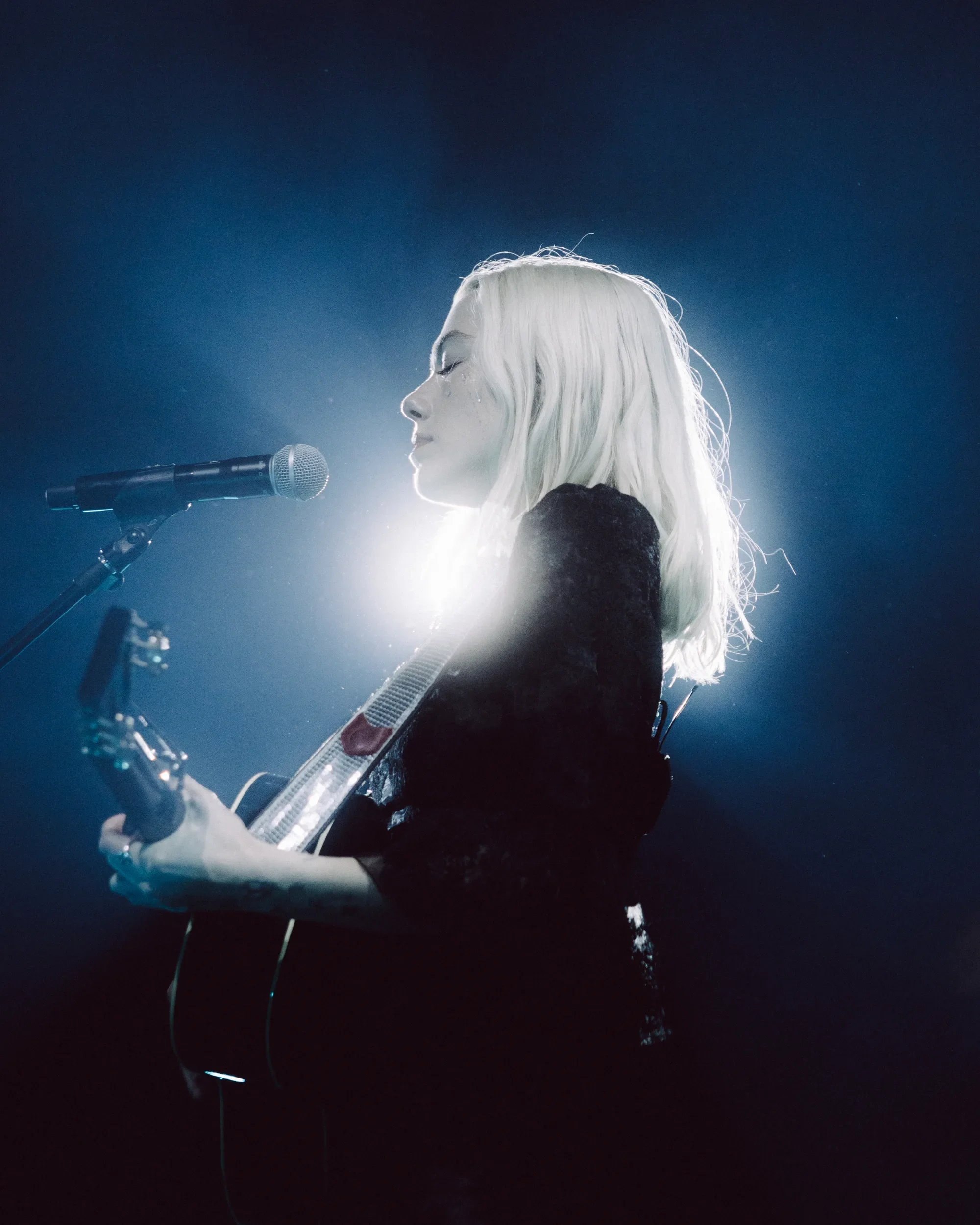 Phoebe Bridgers holds her guitar at a microphone, with bright backlighting behind her at Rogers Arena. Photo: Wes Ellis / @atwes.