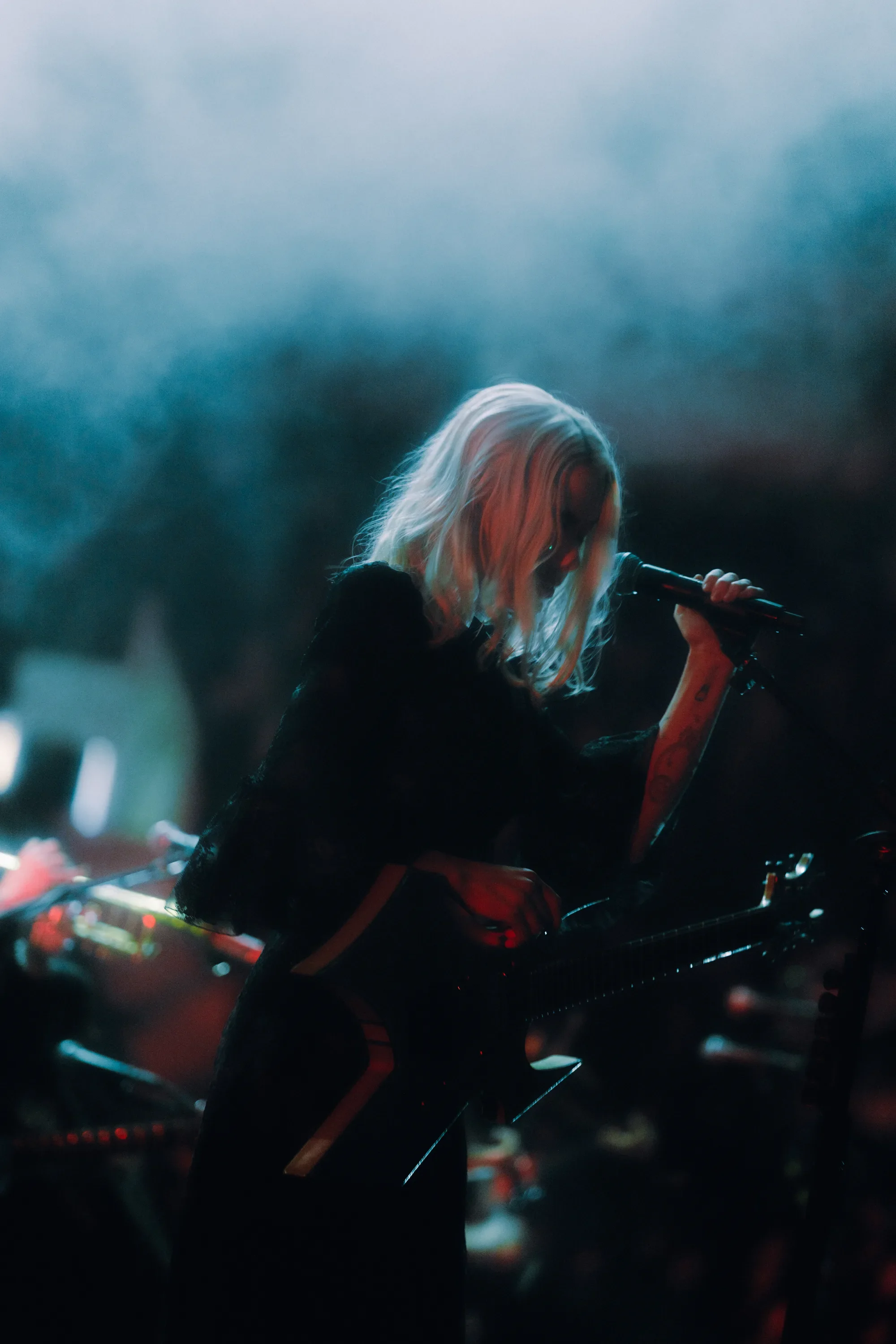 Phoebe Bridgers leans toward a microphone with her guitar beneath blue stage haze at Rogers Arena. Photo: Wes Ellis / @atwes.